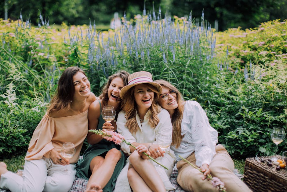 A group of four women sitting on a picnic blanket outdoors in a lush garden near Morden Hall Park, surrounded by tall purple and yellow wildflowers and green foliage under bright natural light. They a
