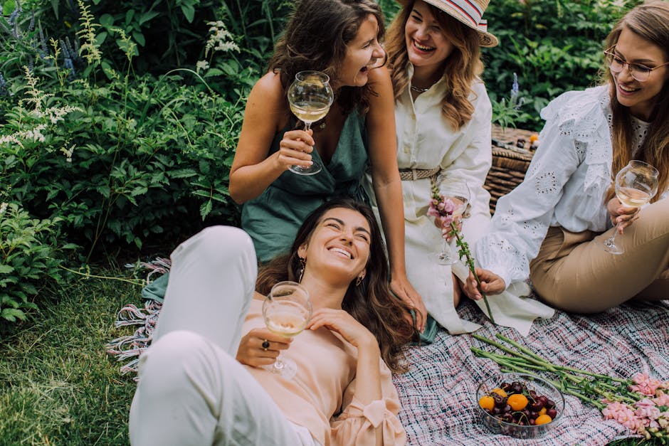 Four women enjoying a picnic outdoors at Morden Hall Park, surrounded by lush greenery and tall white flowering plants. One woman is lying on her back on a patterned blanket, holding a glass of white 