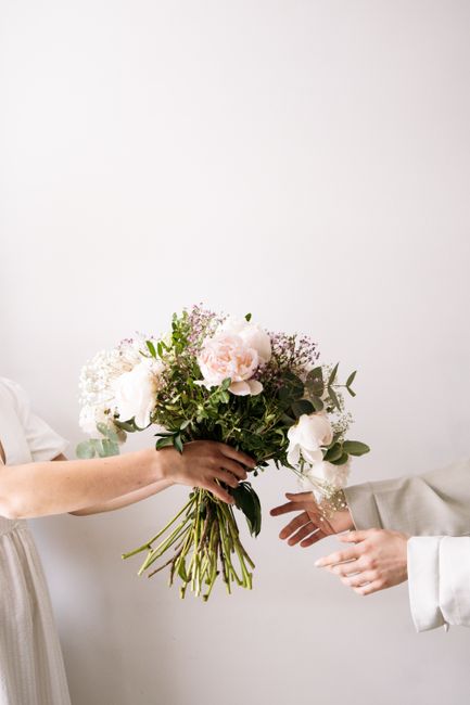 A person in a white dress is holding a large, elegant bouquet of fresh flowers, featuring soft pink peonies, white roses, and sprigs of baby's breath, accented with lush green foliage. The bouquet is