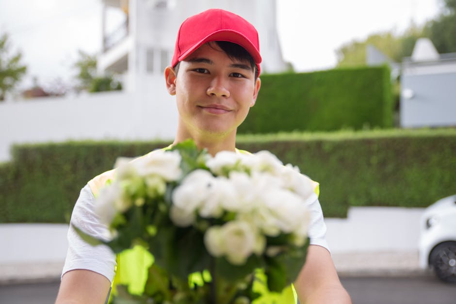 A delivery person wearing a red cap and a high-visibility vest is standing outdoors on a residential street, holding a bouquet of fresh white roses, lilies, and other flowers wrapped in clear cellopha