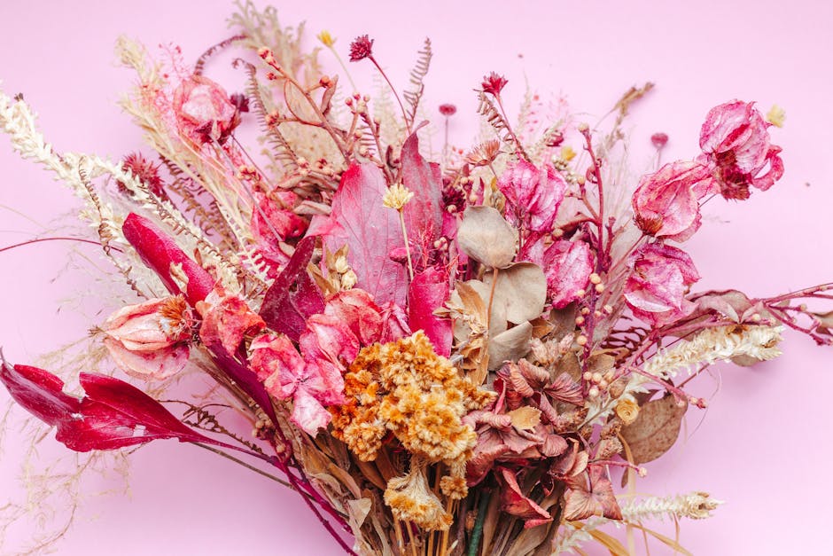 A bouquet of dried flowers resting on a pale pink background, featuring a variety of textures and colors. The arrangement includes faded pink roses, deep red tulip-like blooms, beige and brown leaves,