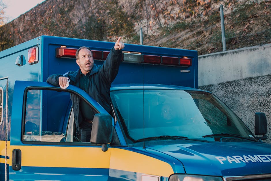 A man wearing a dark jacket is standing outside a blue paramedic emergency vehicle, leaning on the open driver's side door. He has short brown hair and appears to be speaking or giving directions, poi