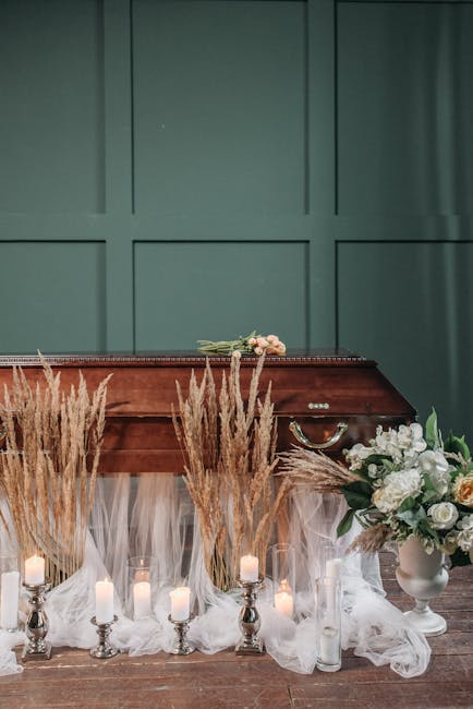 A floral arrangement featuring white roses, peonies, and greenery placed in a white vase on a wooden surface, complemented by an assortment of dried beige pampas grass in front. Surrounding the flower