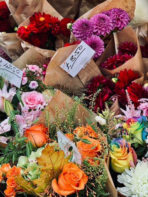 A vibrant assortment of fresh flowers arranged in paper wrapping, including red dahlias and pink chrysanthemums at the top, with a bouquet of orange roses, peach lisianthus, and multicolored roses in 