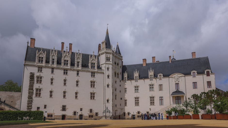 A white historic castle with multiple towers and turrets, featuring ornate gothic windows and decorative stonework, set against a cloudy sky. The castle is surrounded by a paved courtyard with potted 