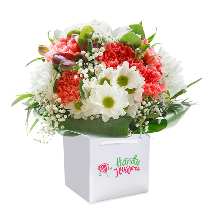 This lush, locally inspired floral arrangement - ideal for delivery across Merton - is beautifully presented in a modern, square white gift bag, staged against a crisp, white backdrop. The bouquet is a striking medley of soft, peachy-coral carnations with ruffled, abundant petals, paired artfully with pristine white daisies that boast vivid, yellow-green centers. Interspersed among these focal flowers are clusters of white baby's breath, lending a gossamer, airy feel, while glossy, broad green leaves create a cradle of foliage at the base, elegantly supporting the blooms and providing a vibrant natural frame. Scattered throughout, subtle purple-tinged buds, possibly alstroemeria, add both depth and a touch of intrigue. The thoughtful composition, rounded and brimming with life, radiates freshness and celebration, much like the vibrant energy found at Merton's local flower markets or community festivities near Wimbledon Common. Sturdy ribbon handles offer easy lifting for same-day delivery or a walk along Colliers Wood with a surprise bouquet in hand. Every detail - from the meticulous placement of daisy petals to the delicate interplay of textures between gypsophila and carnation - speaks of local craftsmanship and care. The clean, uncluttered light focuses attention on the flowers' natural vibrancy and makes the bouquet an ideal gift for marking a joyful milestone, sending congratulations, or simply letting someone in Merton know they're thought of with kindness.