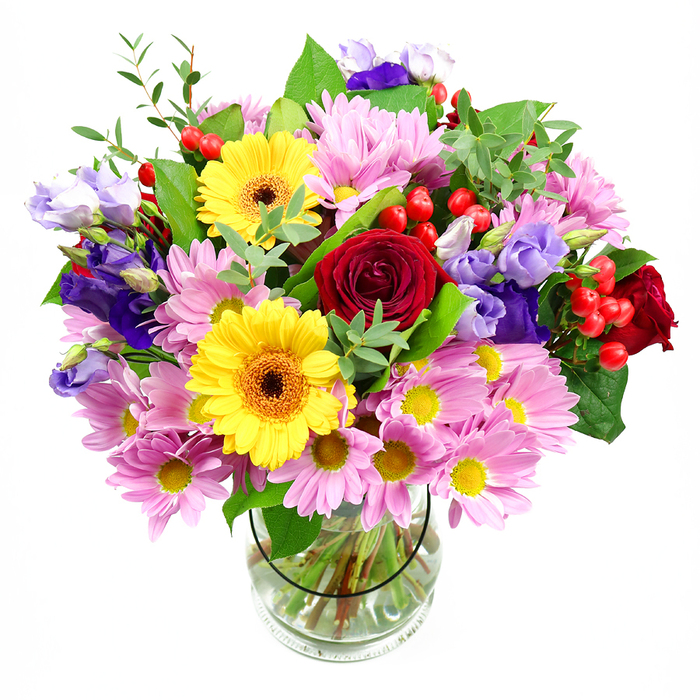 A lush, overflowing bouquet stands proudly in a simple clear glass vase, with a clean white backdrop that highlights every nuance of colour and form. The composition is thoughtfully layered, beginning with a core of deep red roses whose velvety petals curl inward and outward in rich spirals, giving the heart of the design a sense of depth and intimacy. Radiant yellow gerberas, each with a warm brown-orange centre, fan out from this core, their bold, daisy-like faces catching the light and adding a golden, uplifting energy. Encircling these are abundant pastel pink chrysanthemums or daisies, their numerous slender petals and small yellow centres forming a soft collar of texture and gentle hue, like a feminine frame around the stronger blooms. Interspersed among them, slender stems of purple and lavender lisianthus rise and dip, their elegant, bell-shaped flowers bringing a cool, refined note that tempers the brighter tones and introduces a touch of quiet sophistication. Scattered through the foliage, bright red Hypericum berries cluster in small, rounded beads, giving the eye something unexpected to discover, and adding a fresh, berry-like accent. The greenery is varied and generous: broad dark leaves provide structure, while lighter, more delicate sprigs and tips soften the edges, creating an organic, garden-picked feeling. Through the transparent vase you can see the neatly trimmed stems fanning into clear water, a detail that hints at professional care and same-day freshness, as if it has just arrived from a trusted Merton florist on Kingston Road. The mood of the bouquet is joyful yet composed, ideal for celebrations at home near Wimbledon Chase, quiet thank-yous in an office near Morden, or simply brightening a kitchen table with a burst of colour and life.
